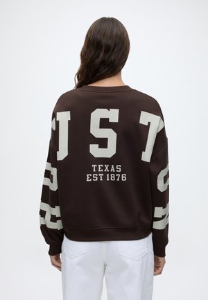 Persoon met lang donker haar die een donkerbruine trui draagt met witte tekst "TEXAS EST 1876" en grote letters op de mouwen en rug.
