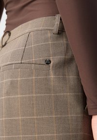 Gros plan sur un pantalon en tissu à carreaux marron avec une poche latérale et une passant de ceinture, porté avec un haut marron à manches longues ajusté.