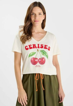 Vrouw met golvend bruin haar, gekleed in een witte T-shirt met V-hals met rode tekst "CERISES" en kersgrafiek, gecombineerd met een olijfgroene rok en oranje trekkoord.
