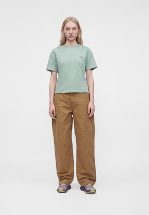 Junge Frau mit langen blonden Haaren, trägt ein hellgrünes kurzärmeliges Hemd, braune lockere Hosen und lilafarbene Sneakers, steht frontal.