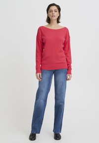 Pull en maille rouge avec un large décolleté et des manches longues, associé à un jean droit bleu et des chaussures noires. Texture lisse et coupe décontractée.