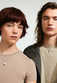 Et par personer iført tætsiddende toppe i neutrale nuancer. Begge bærer sølvhalskæder med rektangulære vedhæng. Baggrunden er lys og minimal.