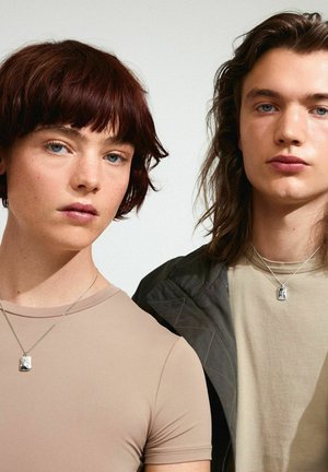 Twee personen in neutrale, getailleerde tops. Beide dragen zilveren kettingen met rechthoekige hangers. De achtergrond is licht en minimalistisch.