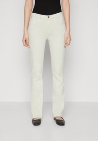 Jean évasé blanc avec une texture lisse, design à cinq poches et un seul bouton marron au niveau de la taille. Porté avec des ballerines noires.
