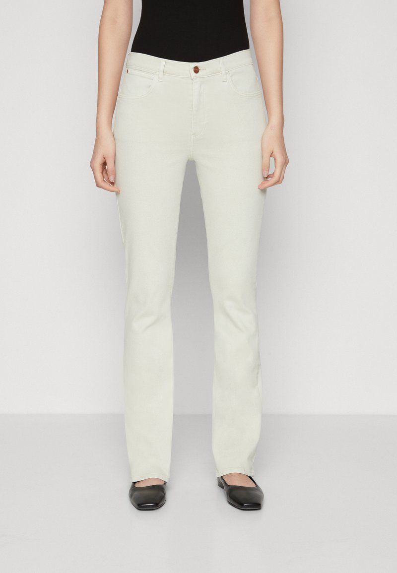 Jean évasé blanc avec une texture lisse, design à cinq poches et un seul bouton marron au niveau de la taille. Porté avec des ballerines noires.