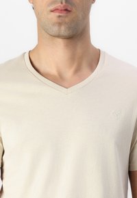 Beige V-Ausschnitt T-Shirt aus weichem Stoff; mit kurzen Ärmeln und einem dezenten gestickten Logo auf der Brust. Glatte Textur.