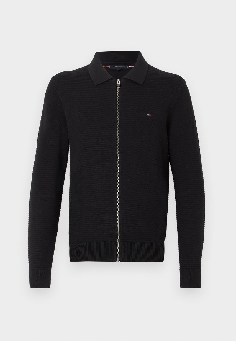 Black knit zip-up sweater with a ribbed collar and cuffs, featuring a small logo patch on the chest. Textured fabric and fitted design.