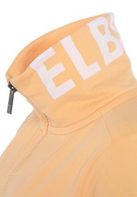 Felpa con zip in cotone arancione pastello con colletto alto. Presenta testo bianco lungo il colletto e una superficie liscia.