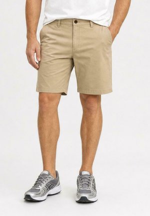 Man draagt beige knielange shorts, wit T-shirt en grijze hardloopschoenen, staat met handen in de zakken.