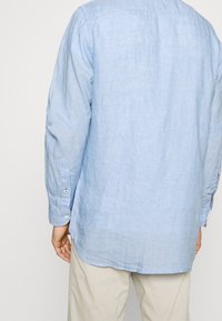 Chemise boutonnée à manches longues de couleur bleu clair, fabriquée en lin. Présente un ourlet arrondi et des détails de couture visibles aux poignets.