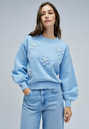 Pull en tricot bleu clair avec des manches bouffantes et des poignets côtelés, orné de fleurs blanches brodées et d'un col rond, associé à un jean bleu clair.