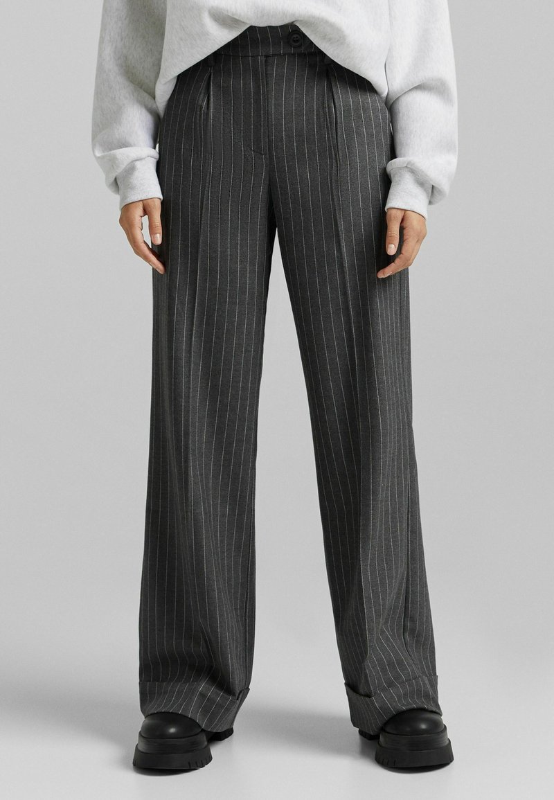Personne portant un pantalon large à fines rayures gris foncé avec des ourlets retroussés, des chaussures noires épaisses, et un pull oversize gris clair.