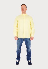 Camicia button-up giallo chiaro con maniche lunghe, indossata su jeans in denim blu e abbinata a sneaker blu scuro.