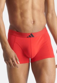 Röda boxershorts i stretchmaterial med en svart midja som har Adidas-loggan. Designen inkluderar en stödjande fack och en slät yta.