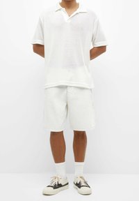 Camisa polo de malla blanca con cuello corto, combinada con pantalones cortos de mezclilla de color claro. Calcetines tobilleros blancos y zapatillas con acentos negros completan el atuendo.