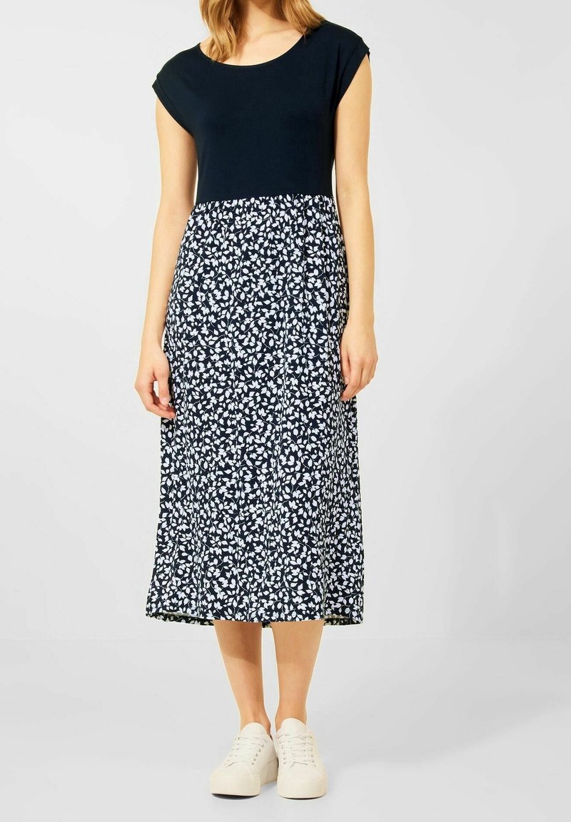 Granatowy top z rękawkami typu kepi łączy się z floralnie wzorzystą spódnicą do kolan w kolorach białym i niebieskim. Noszony z białymi trampkami. Gładka faktura tkaniny.