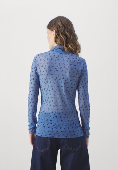 Top lungo a maniche trasparenti blu con motivo floreale in tonalità viola, colletto alto e vestibilità aderente, abbinato a jeans in denim scuro.