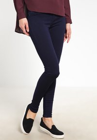 Jeans ajustados de color azul marino, confeccionados en denim elástico, combinados con zapatillas negras tipo slip-on. Los jeans cuentan con un clásico diseño de cinco bolsillos.