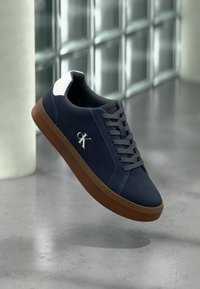 Baskets Calvin Klein bleu marine avec semelle marron, lacets gris et détail blanc au talon, flottant au-dessus d'un sol en béton poli.