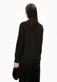 Schwarzer Pullover aus weichem Material, mit lässigem Schnitt, langen Ärmeln und gerippten Bündchen, kombiniert mit dunklen Cordhosen und einem dunklen Accessoire.