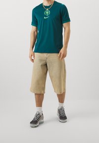 Camiseta azul verdoso de Nike con logo bordado, combinada con pantalones cortos cargo beige claro. Zapatillas grises y negras con suela visible de aire.