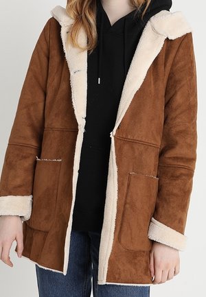 Personne portant une veste en daim marron avec une doublure en shearling blanc sur un sweat à capuche noir et un jean bleu, mains détendues sur les côtés.