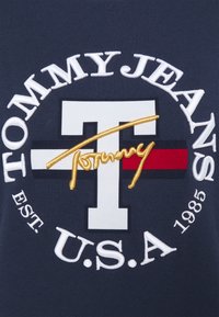 Logo Tommy Jeans avec un grand T blanc, des rayures bleu marine, rouge et noir, le script doré "Tommy" et le texte blanc "Tommy Jeans U.S.A Est. 1985" sur un tissu bleu marine.