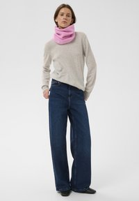 Világosszürke kötött pulóver, széles szabású kék farmernadrág, és bordázott rózsaszín kötött sál. A modell zsebre tett kézzel áll, tiszta háttér előtt.