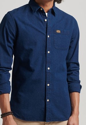 Man draagt een donkerblauw hemd met lange mouwen, een borstzak en opgerolde mouwen, zichtbare beige broek bij de taille.