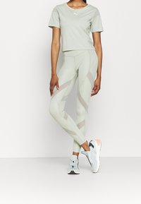 T-shirt court vert clair avec un petit logo, assorti à des leggings verts clairs ornés de panneaux transparents et de motifs géométriques. Baskets grises et roses.