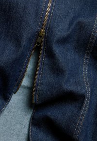 Tumma sininen denim-kangas, jossa on kultainen vetoketju, näkyviä ompeleita ja pehmeä sisävuori. Tekstuurissa on hieman ryppyjä.