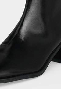 Gros plan d'une botte en cuir noir brillant avec un bout pointu et un petit talon bloc sur fond blanc.