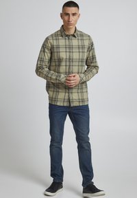 Beige ternet skjorte med sorte linjer, knaplukning, lange ærmer og klassisk krave, parret med mørkeblå jeans og sorte sneakers.