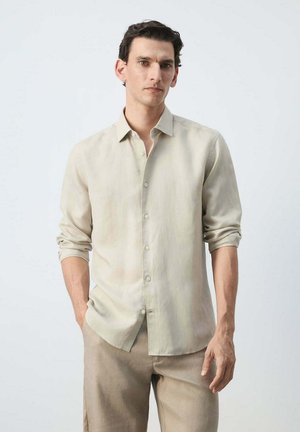 Mand med mørkt krøllet hår iført en beige skjorte med knapper og opkørte ærmer samt matchende beige bukser mod en ensfarvet lys baggrund.