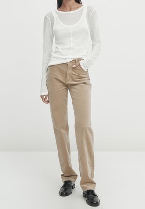 Femme portant un haut blanc à manches longues semi-transparent, un pantalon beige droit et des chaussures en cuir noires à lacets, debout avec une main dans la poche.