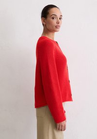 Czerwony sweter z dzianiny z długimi rękawami, okrągłym dekoltem i guzikami z przodu. Teksturowany wzór i lekko luźny krój; zestawiony z beżowymi spodniami.