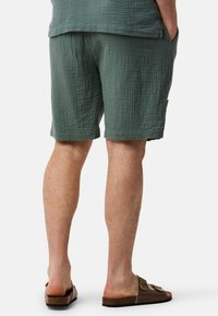 Shorts verts texturés avec une coupe décontractée, présentant des poches latérales et un ourlet simple. Portés avec des sandales à glissement marron.