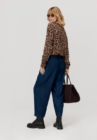 Kobieta w bluzce w leopardzie, ciemnoniebieskich szerokich spodniach, czarnych wiązanych botkach, okularach przeciwsłonecznych i trzymająca dużą ciemnobrązową torebkę.