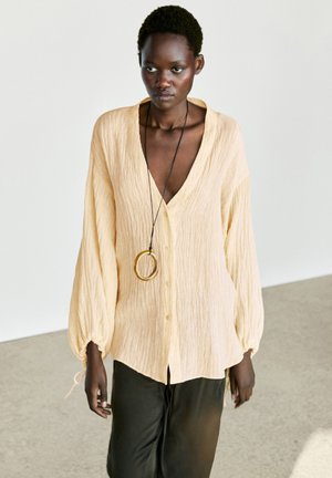 Jeune femme à la peau foncée portant un chemisier ample beige texturé avec des boutons et un long collier avec un grand pendentif circulaire.