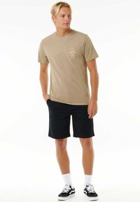 T-shirt in cotone marrone chiaro con un piccolo logo grafico sul petto, abbinata a pantaloni shorts neri e scarpe da ginnastica nere con dettagli bianchi.