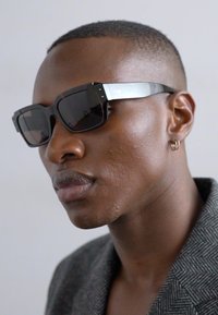 Ochelari de soare negri cu rame pătrate, accente argintii reflective, lentile întunecate, brațe texturate și un detaliu subtil al logo-ului. Purtașul are părul scurt.