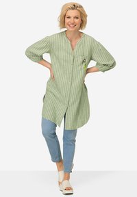 Camicia lunga a righe verdi con bottoni davanti, tasca con dettaglio decorativo, abbinata a jeans azzurri chiari e sandali crema.