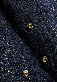 Middengrijze textuurstof met metallic garen, voorzien van gouden accenten en kleine, rozenvormige knopen. Glanzend oppervlak met een ingewikkelde weving.