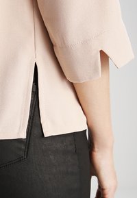 Blusa rosa chiaro a maniche corte con tessuto strutturato, spacchi laterali e un delicato dettaglio sull'orlo, abbinata a pantaloni neri.