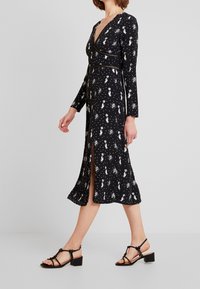 Černé dlouhé šaty s v-výstřihem, květinovým potiskem v bílém, bočním rozparkem a obtaženým pasem. Modelka doplnila outfit o černé sandály na tenkých podpatcích.