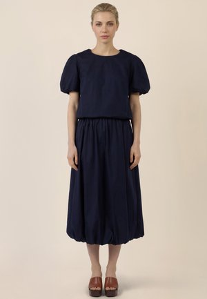 Frau steht frontal und trägt ein marineblaues Oberteil mit Puffärmeln sowie einen dazu passenden, gerafften Midirock und braune Plateausandalen.