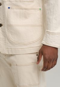 Κοντινή λήψη από τζάκετ από μπεζ υφασμένο ύφασμα· διαθέτει πολλές τσέπες με αντίθετο ραφές και χρωματιστές μεταλλικές λεπτομέρειες σε πράσινο και μπλε.