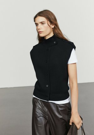 Giovane donna con capelli lunghi indossa un gilet nero abbottonato sopra una camicia bianca e pantaloni in pelle marrone scuro, tenendo una borsa scura nella mano destra.