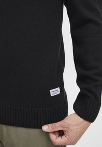Schwarzer Strickpullover mit gerippten Bündchen und Saum, mit einem Etikett am unteren Rand, das Markeninformationen zeigt. Strukturierte Oberfläche.