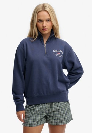 Jeune femme aux longs cheveux blonds portant un sweat-shirt bleu marine à fermeture éclair quart et un short à carreaux verts, debout devant un fond blanc.